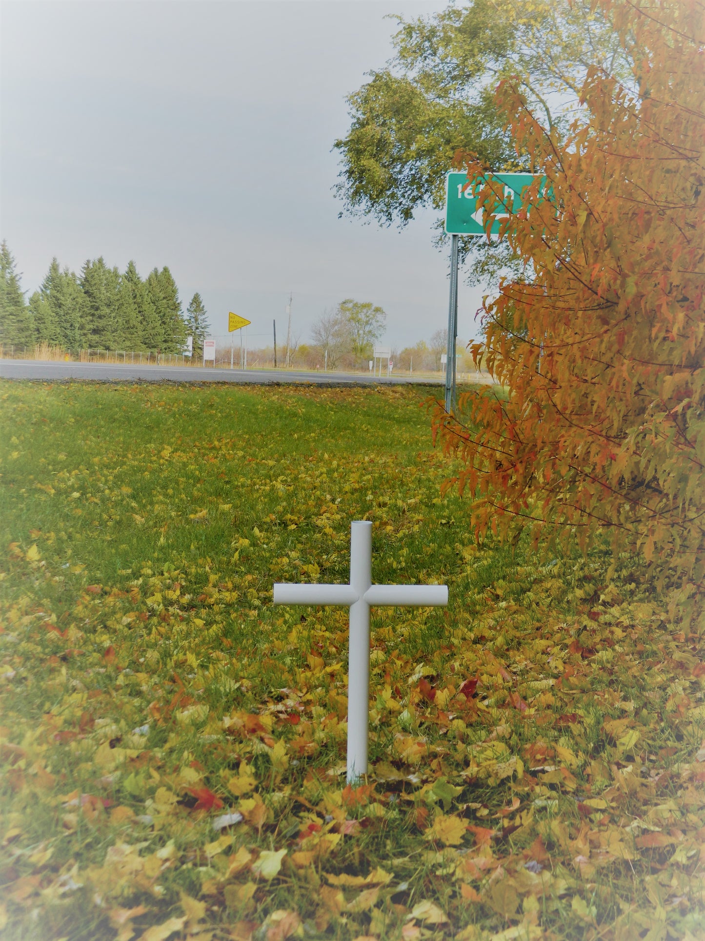 Outside Roadside Memorial Is Timeless With Everlasting Cross. White Finish Baked Powder Coated Over Stainless Steel. It Is Maintenance Free. As It Stands Tall In Its Fall Background Of Oranges, Yellow Leaves located On A Roadside. 